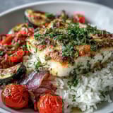 Tender pan-seared fish bowl over jasmine rice, topped with colorful roasted zucchini and bell peppers on a marble counter.