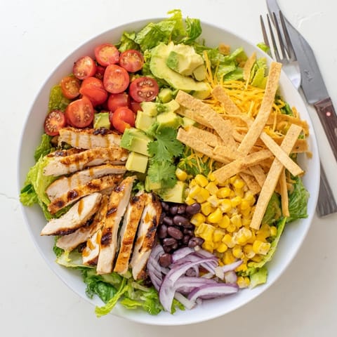 A juicy, charred honey lime chicken breast rests on a bed of mixed greens, sweet corn, black beans, and cherry tomatoes, garnished with red onion and shredded cheese. The salad is ready to serve with a side of lime wedges for extra zest.